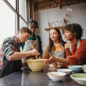 friends preparing food on table while standing against windows in cooking class